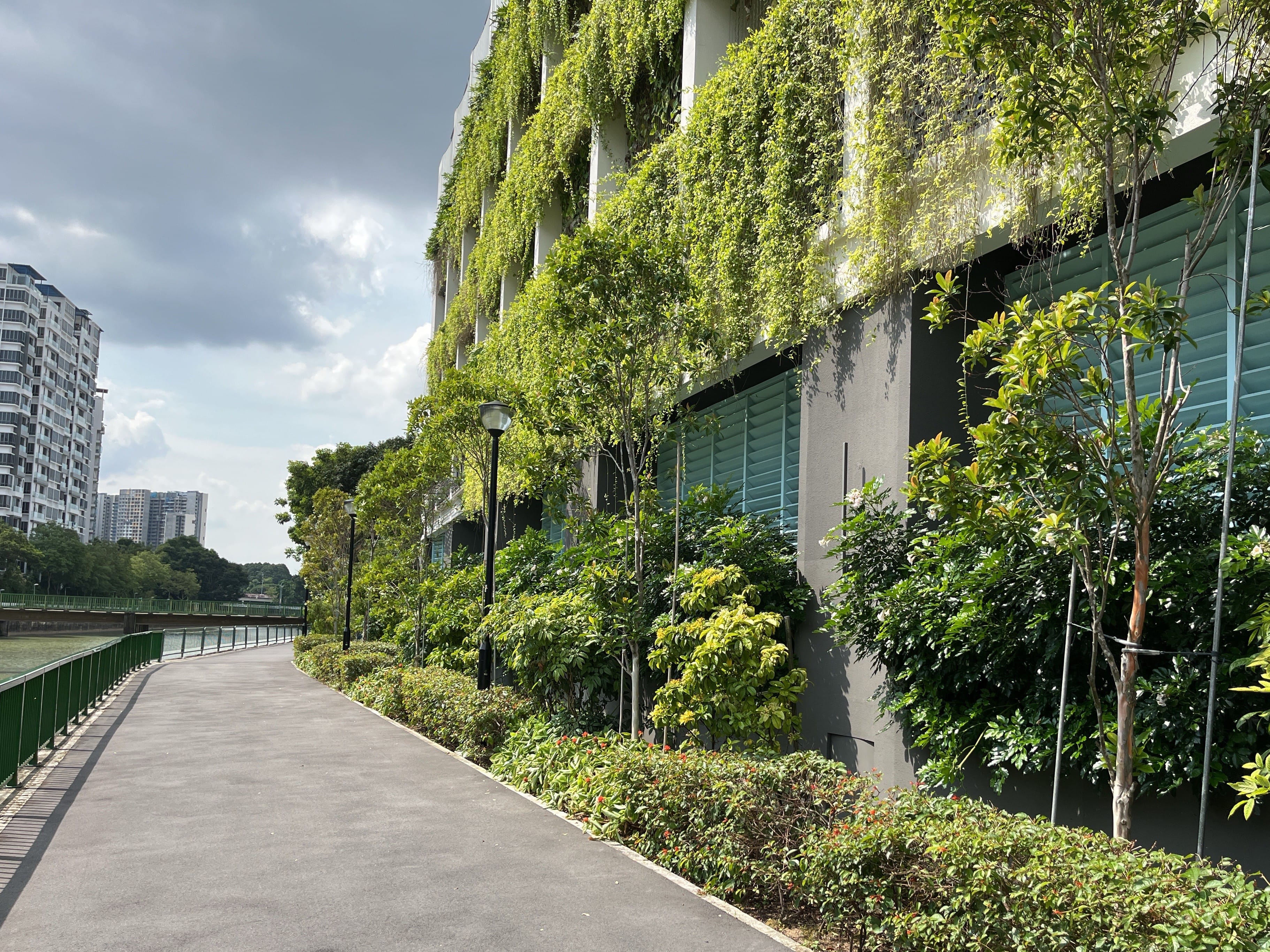 A riverside path beside a building covered with green plants and trees under a cloudy sky.